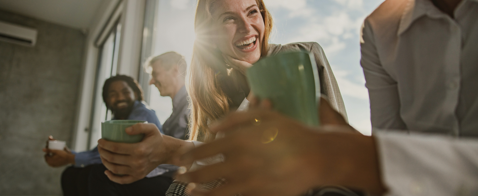 Group of people laughing and holding mugs near a large window with sunlight streaming in.