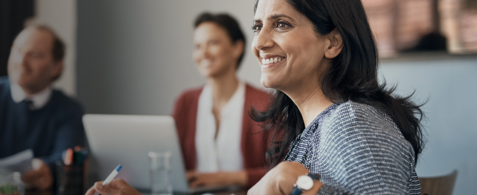 Smiling woman at a meeting table with two blurred colleagues in the background.
